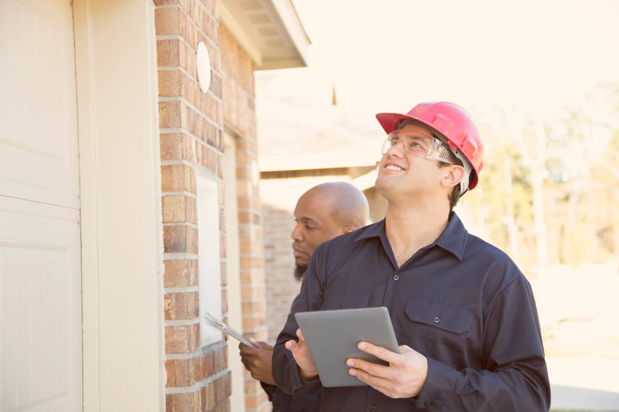 Two property maintenance professionals inspecting a residential building, one holding a tablet and wearing a pink hard hat, emphasizing proactive maintenance strategies in property management.