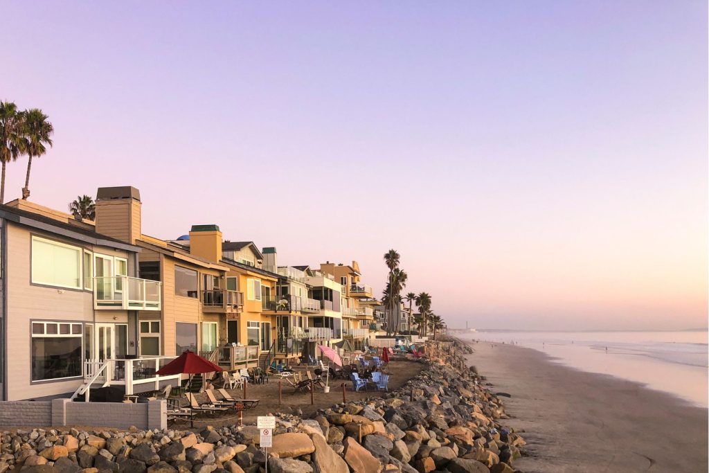 Coastal residential properties along a sandy beach, showcasing modern architecture with balconies, palm trees, and a serene sunset sky, emphasizing community living and property value enhancement.