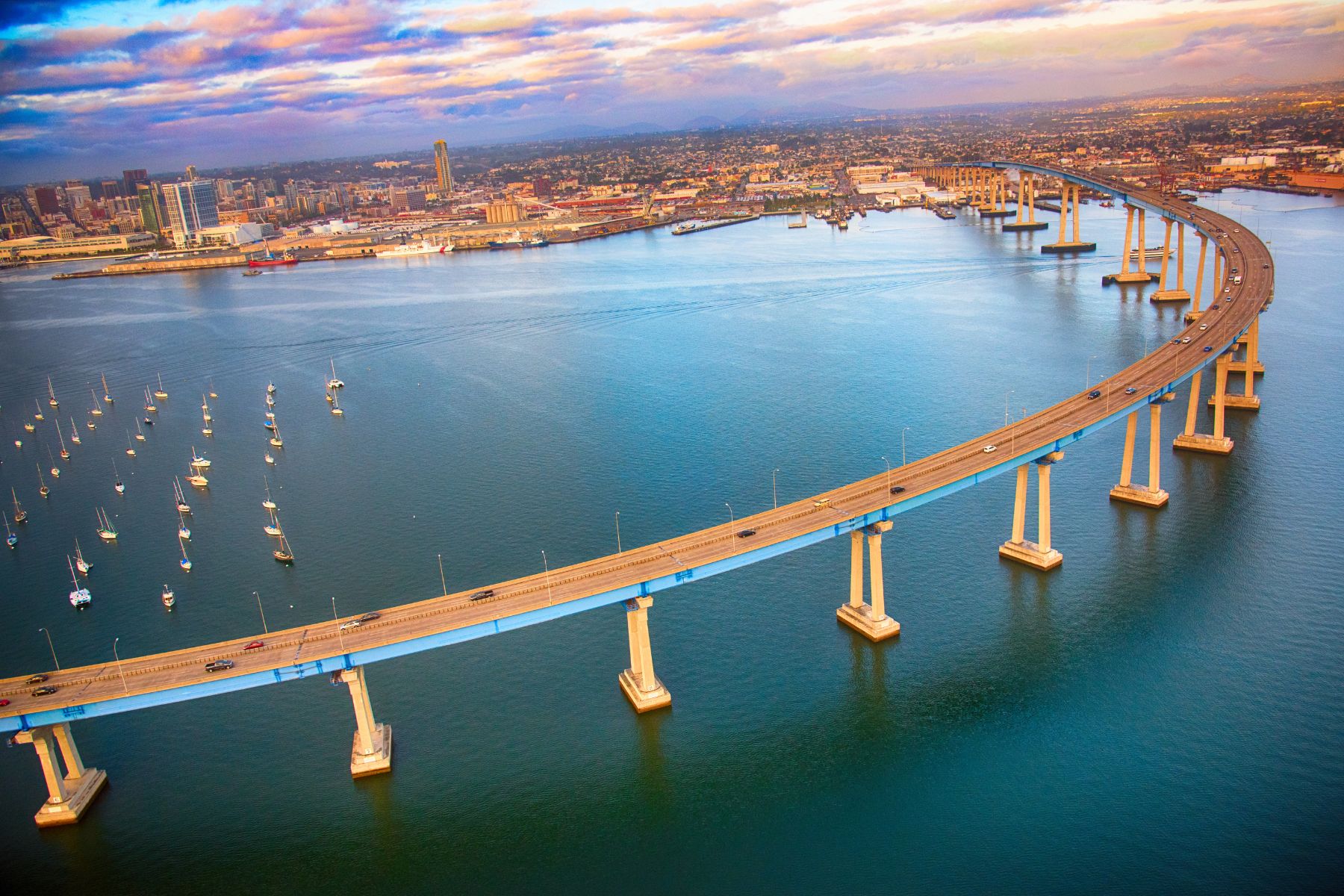 Aerial view of the San Diego-Coronado Bridge spanning blue waters, with sailboats in the foreground and the San Diego skyline in the background, representing coastal property management and investment opportunities in Southern California.