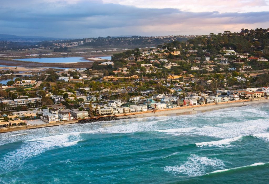Coastal view of a residential community along the shoreline, highlighting properties and amenities in a homeowners association setting, with ocean waves and a backdrop of hills.