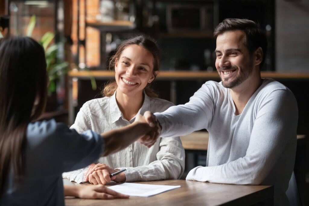 happy smiling millennial couple handshake get acquainted with female real estate agent meeting together in cafe excited husband and wife shake hand of broker or banker thanking for help