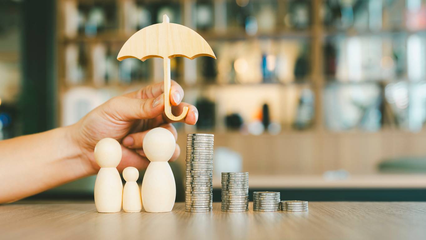 Hand holding a wooden umbrella over a family of three wooden figures and stacks of coins, symbolizing financial protection and security in the context of health and home insurance against rental scams.